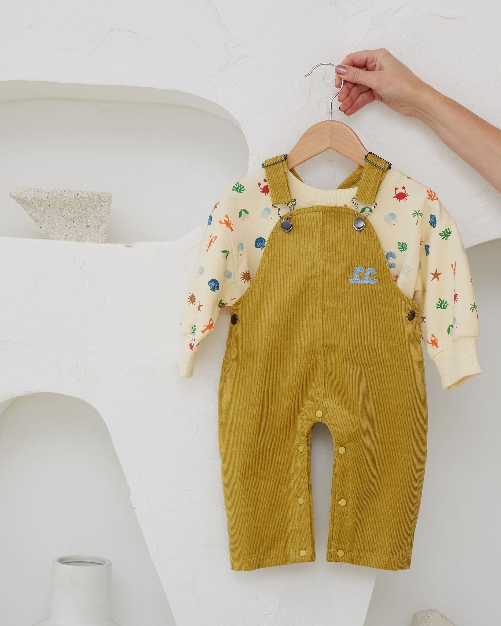 Yellow corduroy overalls with a colorful shirt underneath on a hanger against a white background.